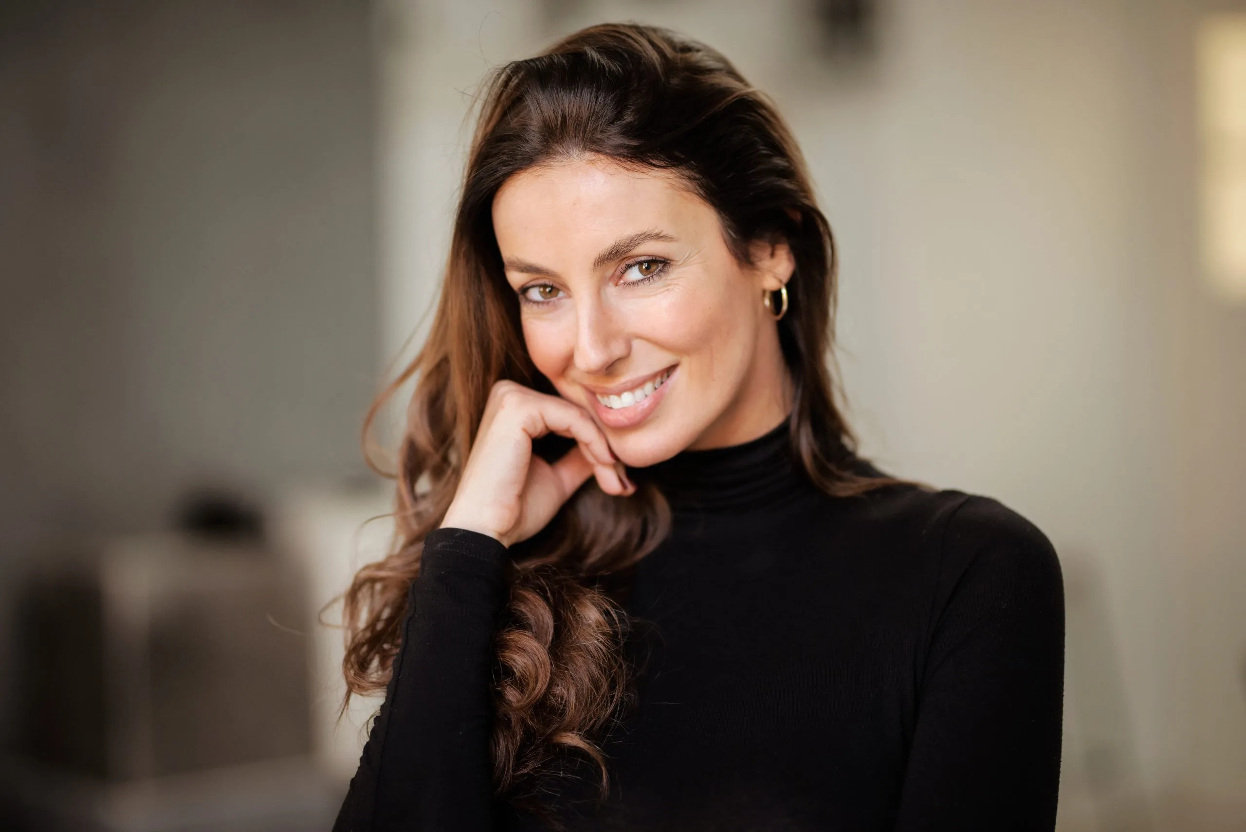 Woman with long brown hair smiling with hand near face against blurred background
