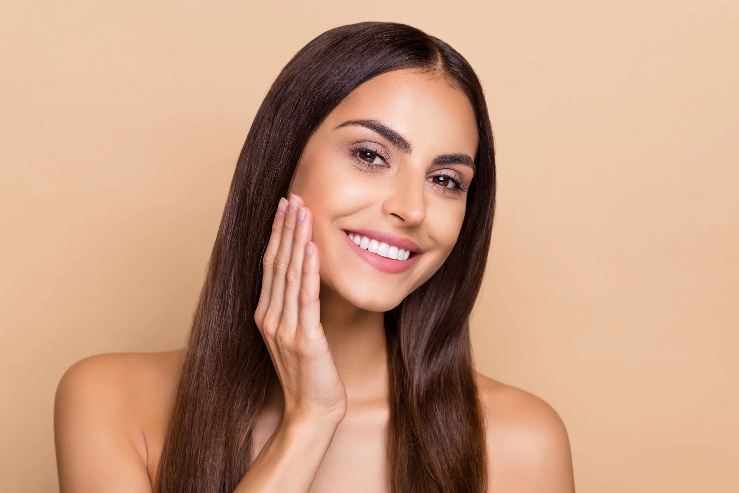 Woman with long dark hair smiling at camera, hand near face showing radiant skin