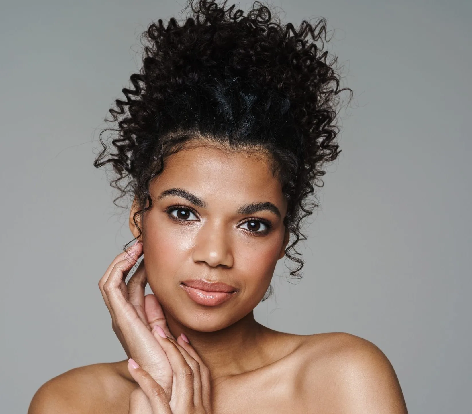 Woman with black curly upswept hair touching face against gray background