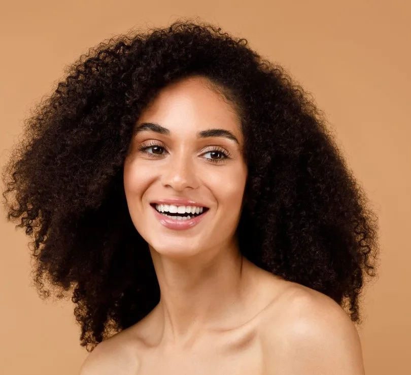 Woman with curly dark hair smiling at camera against tan background