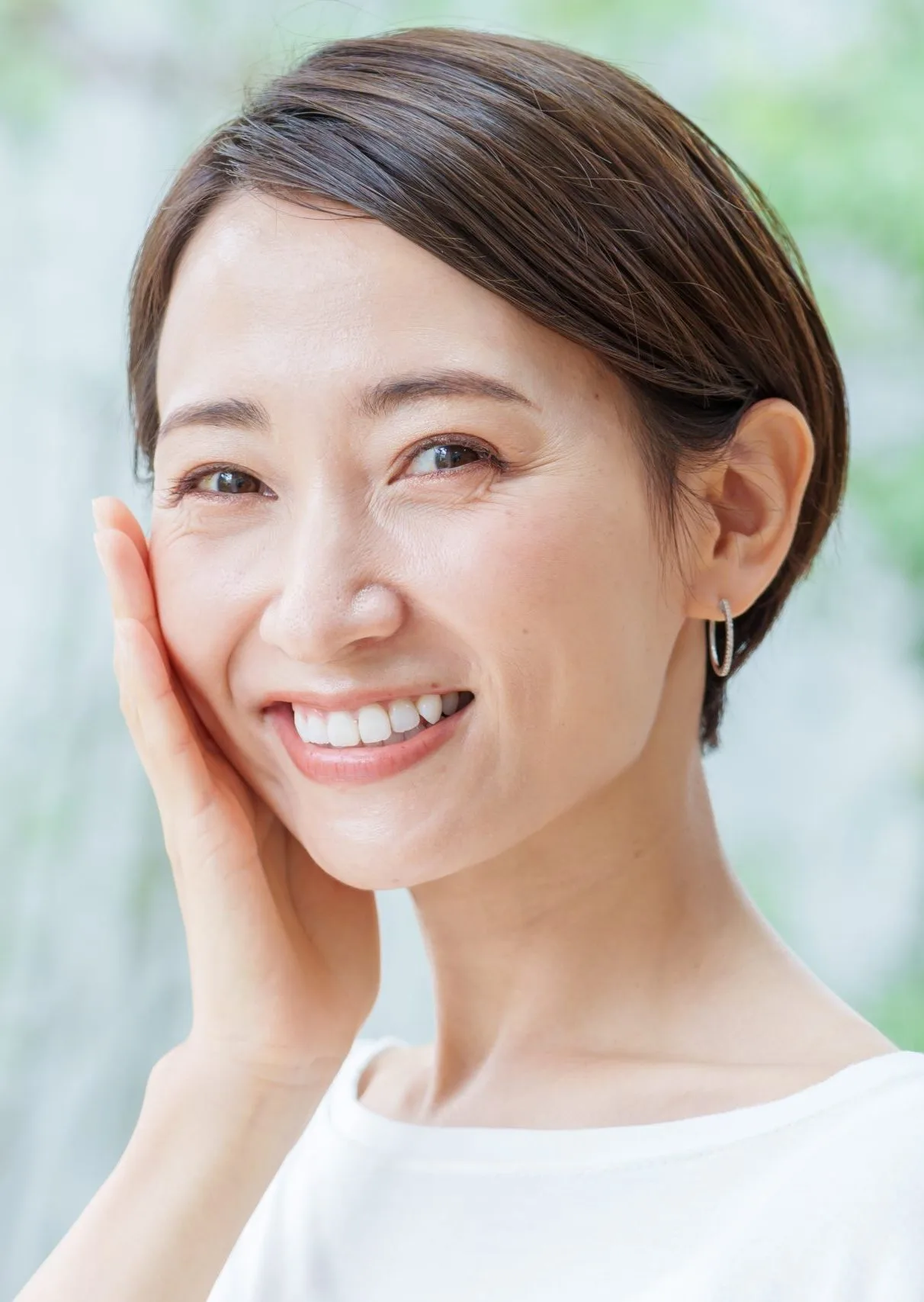Woman with brown hair smiling and touching her face against blurred green background