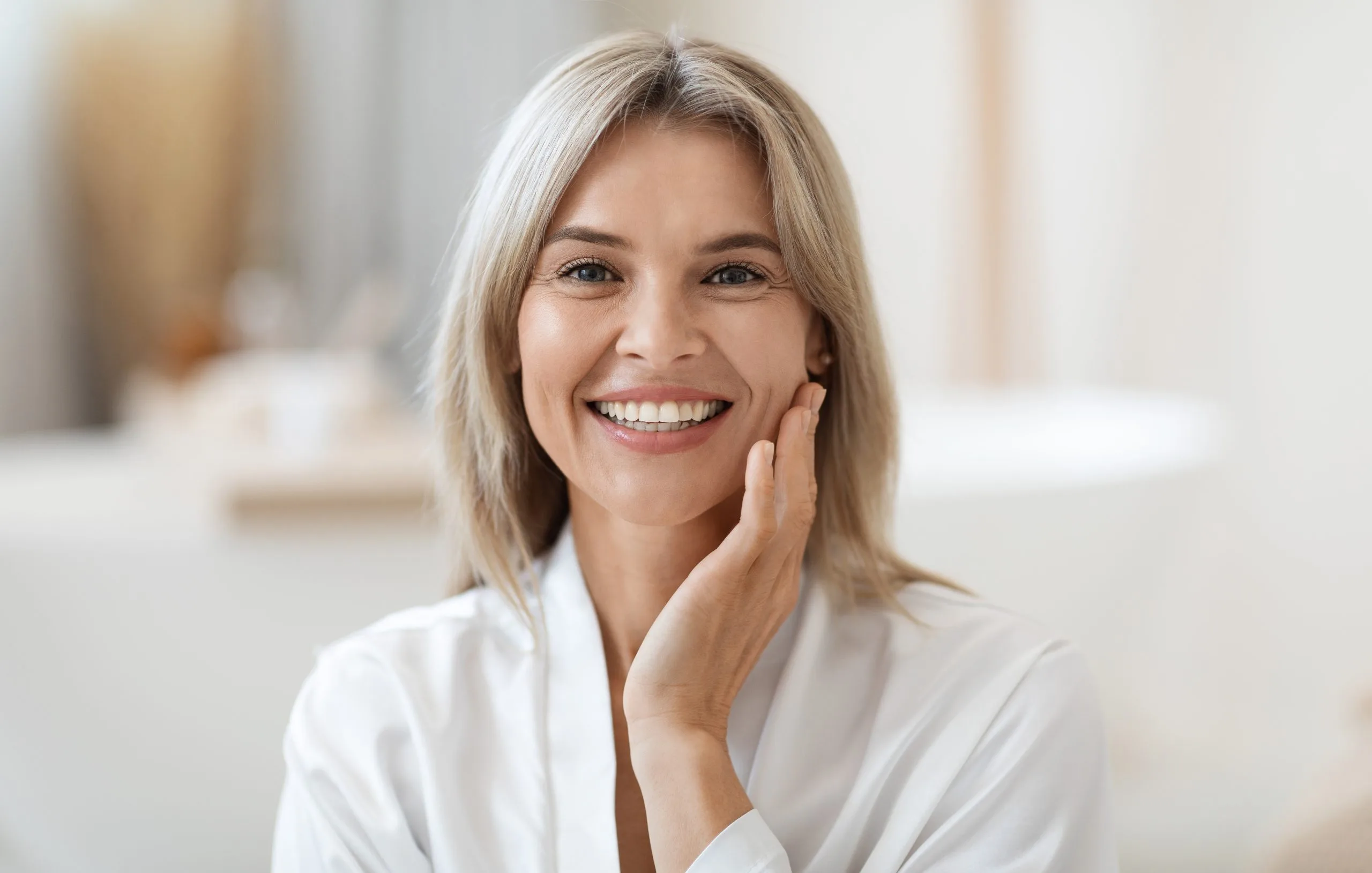 Woman with blonde hair in white shirt touching her face, smiling in a bright indoor setting