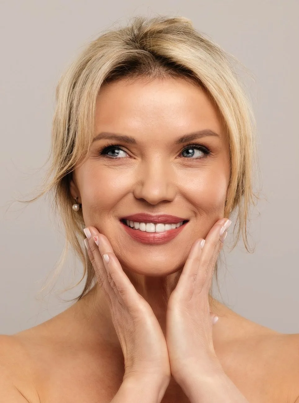 Woman with blonde hair touching her cheeks with both hands, smiling against a light background