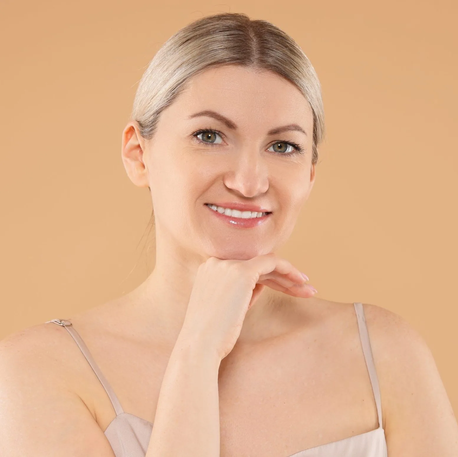 Woman with blonde hair resting chin on hand, smiling at camera against tan background