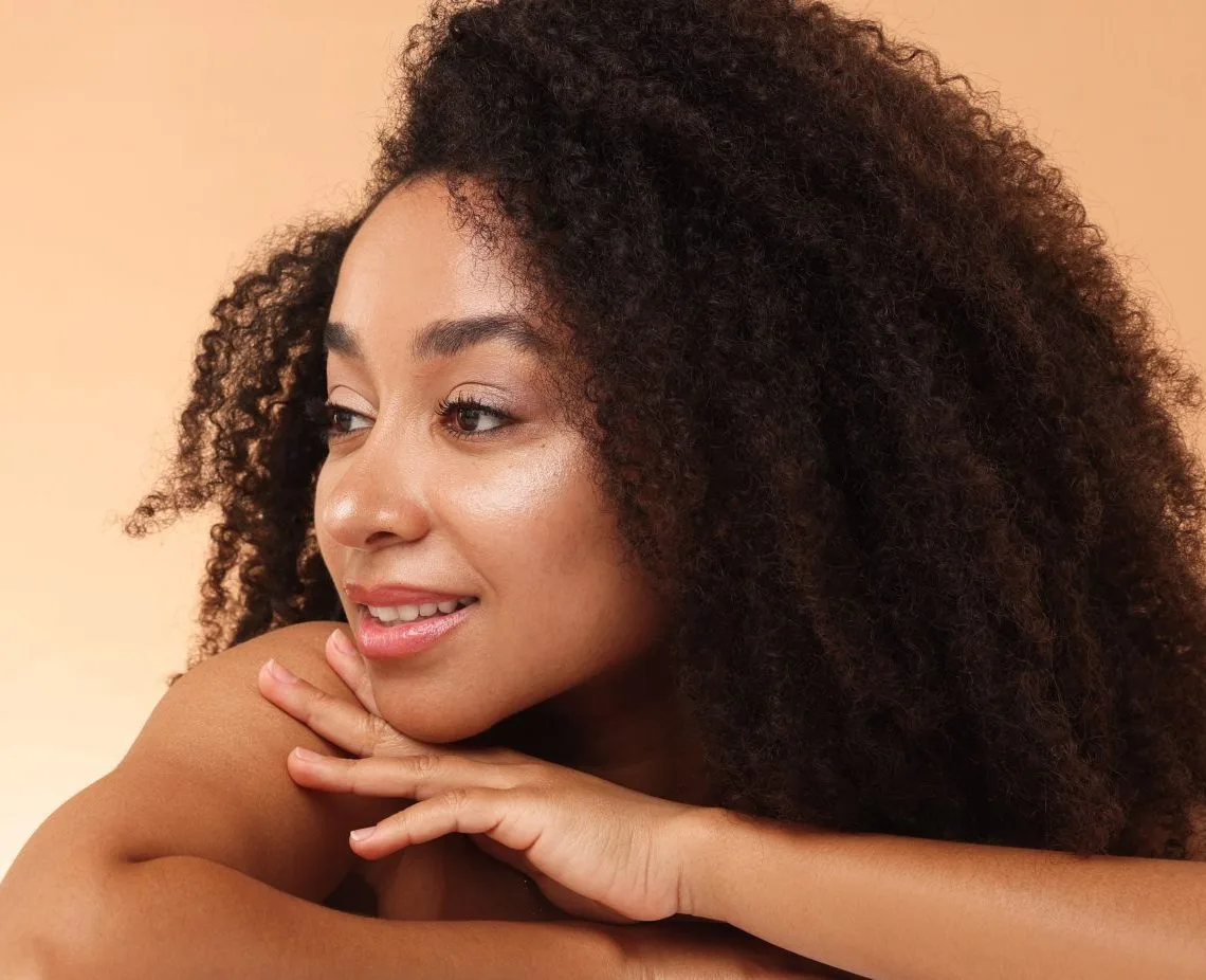 Woman with curly dark hair touching shoulder and smiling against tan background