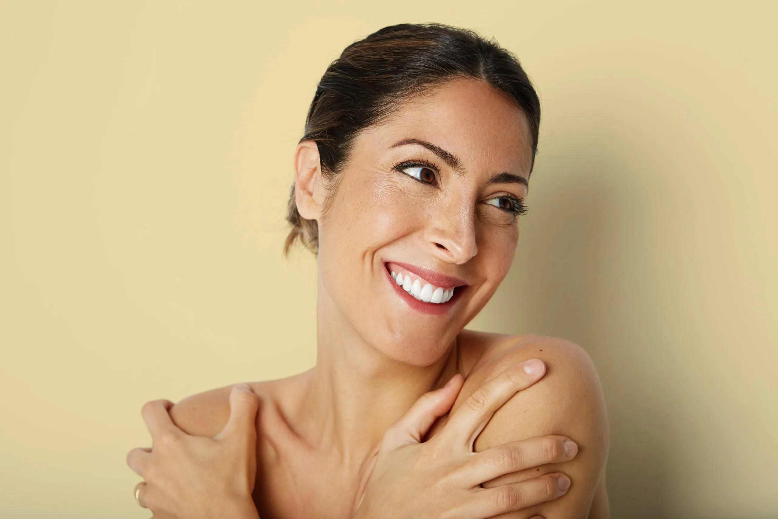Woman with dark hair smiling and touching her shoulder against a yellow background