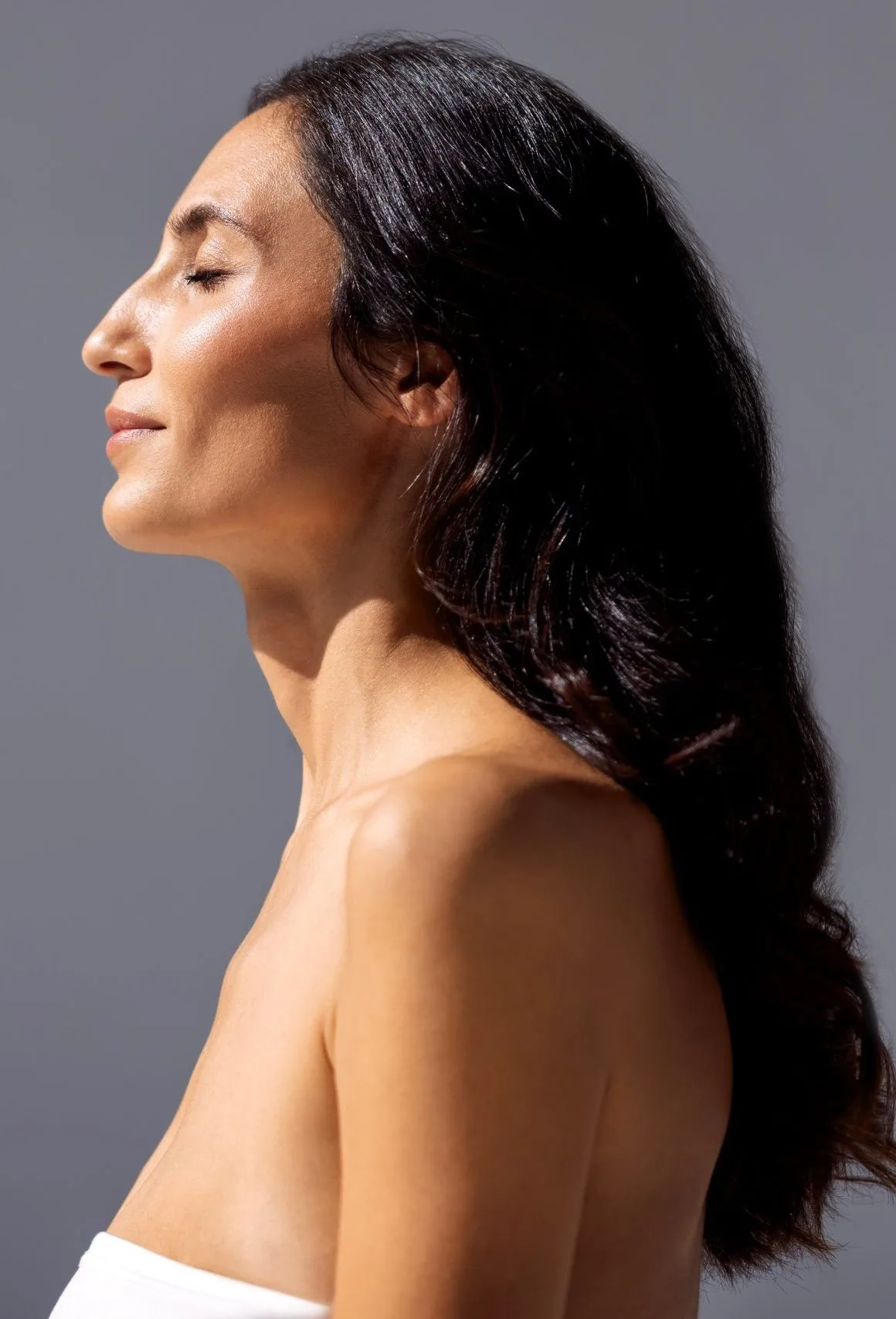 Woman with long dark hair in profile with eyes closed against a gray background