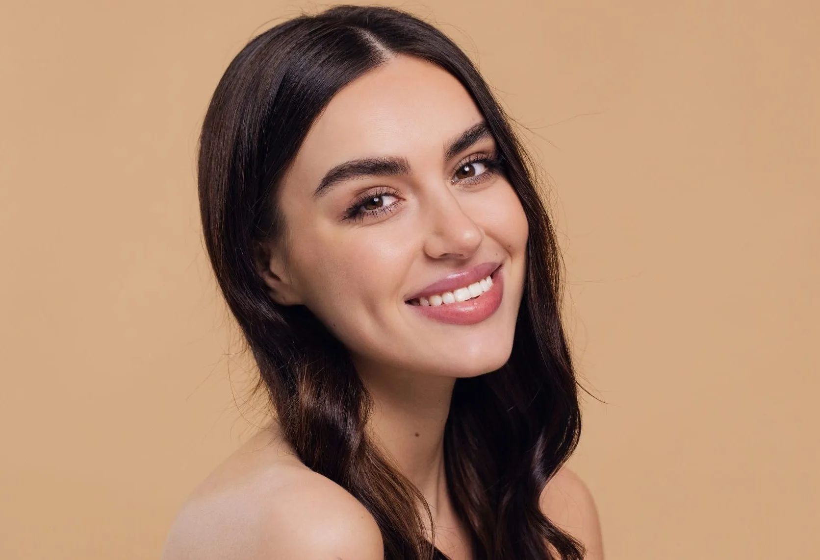 Young woman with clear skin touching her shoulder against a beige background