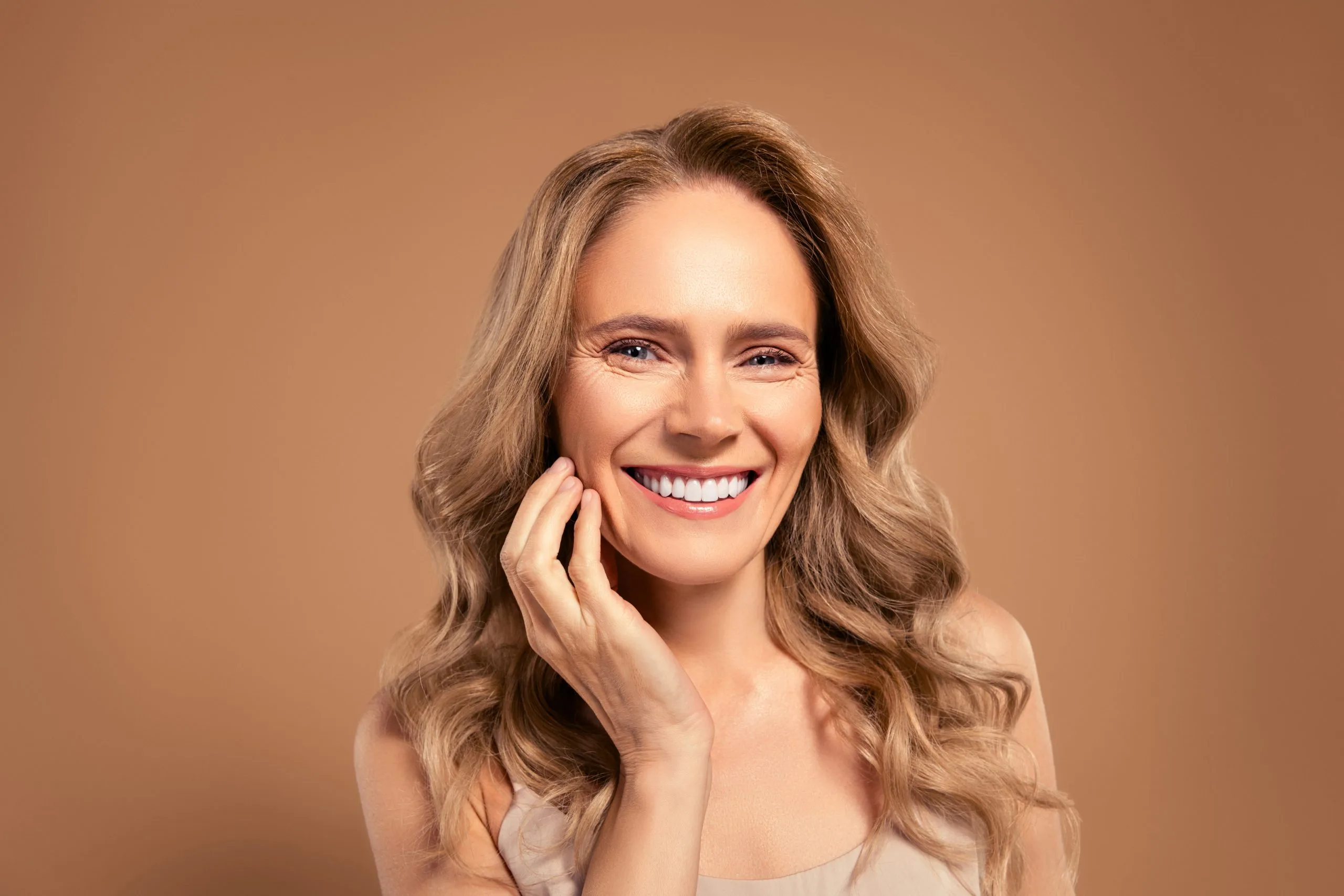 Mature woman with long brown wavy hair smiling confidently against a warm neutral background