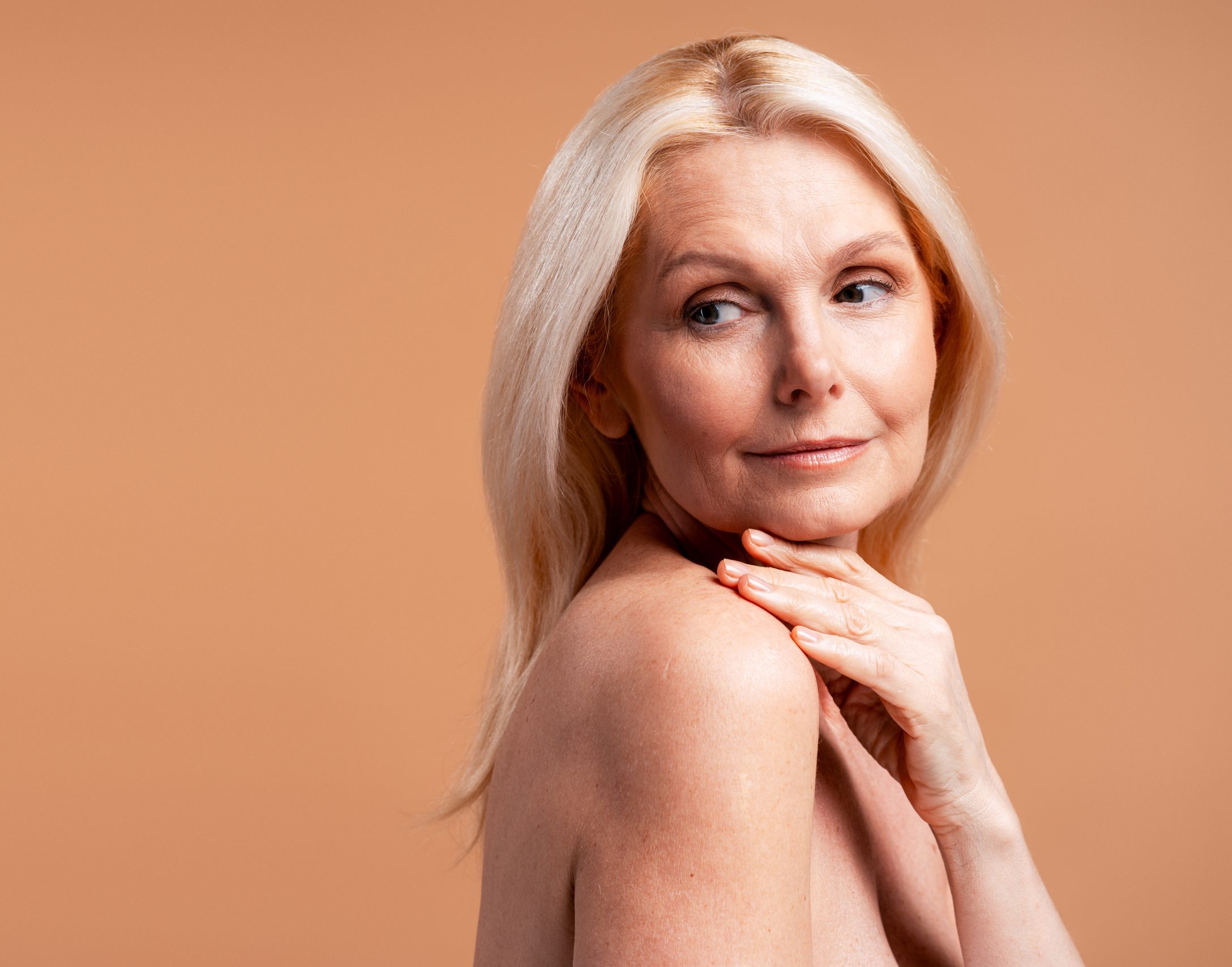Mature woman looking over her shoulder, touching her bare shoulder against a warm peachy background