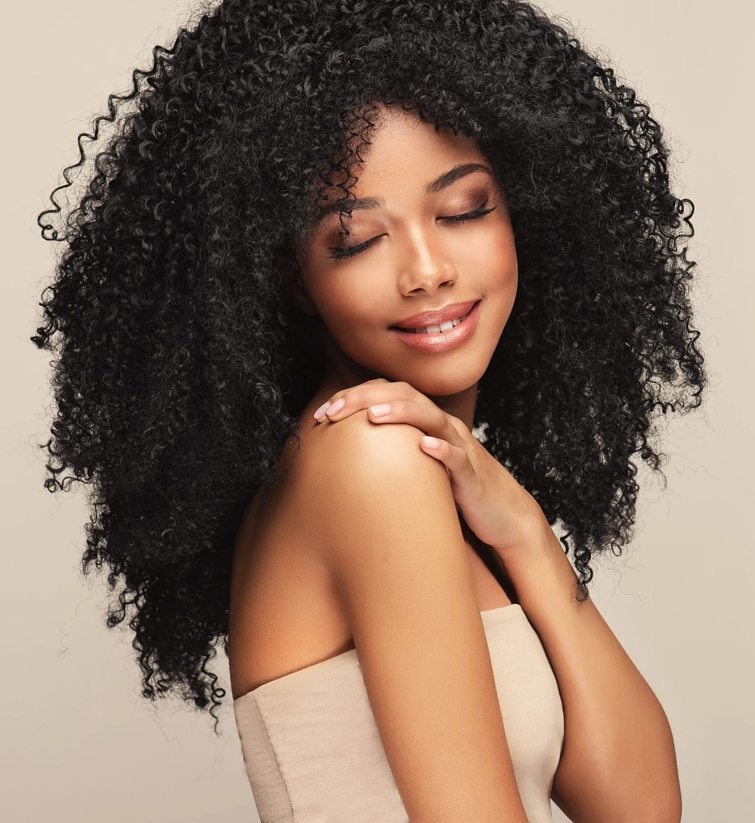 Woman with curly black hair touching her shoulder, displaying smooth skin against neutral background